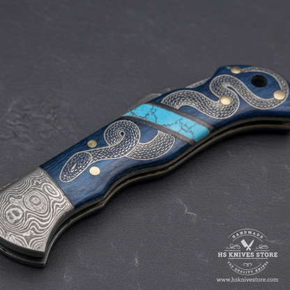 Close-up macro of blue wood handle with hand-engraved silver coiled snake artwork turquoise stone inlay strip and brass dot rivets on Damascus folding knife