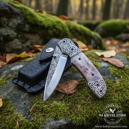 Camel bone handle folding knife open blade resting on mossy rocks in outdoor forest setting with black leather sheath