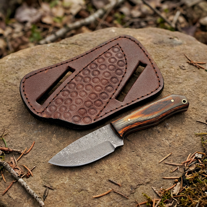 Left-angle view of compact Damascus knife and separate brown sheath on woodland rock