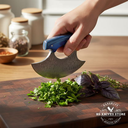 One hand gripping Damascus ulu knife through blue pakkawood handle finger loop in rocking chop motion over dark wood cutting board with herbs