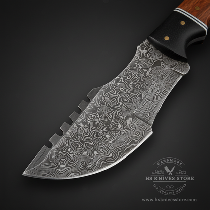 Close-up macro of Damascus steel tracker blade showing bold swirling topographic Damascus pattern and 4 serrated spine teeth and thick spine