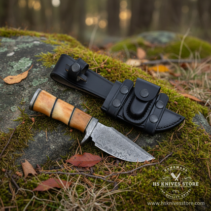 Damascus steel skinner knife camel bone handle resting on mossy rocks in outdoor forest setting with black leather sheath