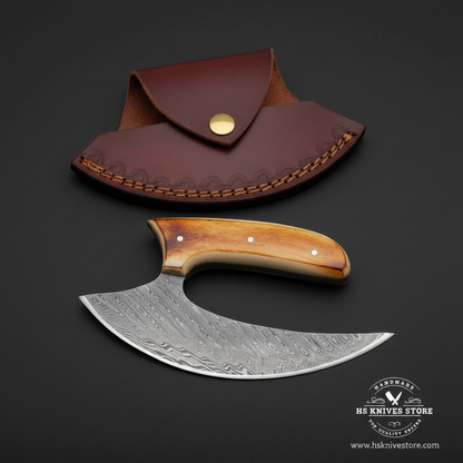 Damascus ulu knife with amber bone handle steel bolster and burgundy crescent leather sheath on dark charcoal studio background facing right