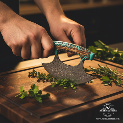 Hands using raindrop Damascus steel ulu knife with pine cone teal resin handle in rocking chopping motion on dark wooden cutting board lifestyle shot
