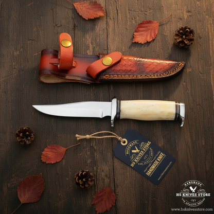 J2 steel hunting knife flat lay with bone handle and HS Knives Store hang tag on rustic dark wood with autumn leaves