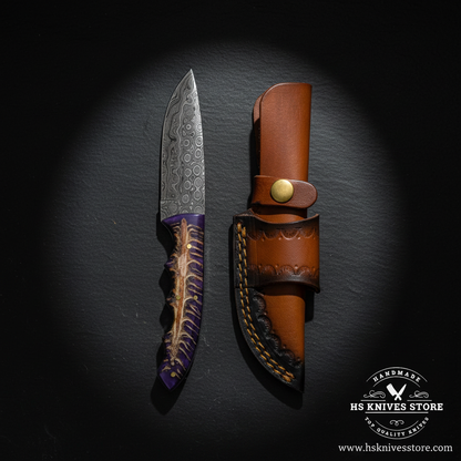 Damascus steel knife displayed vertically on dark slate with purple pine cone stag handle and tan leather sheath luxury shot