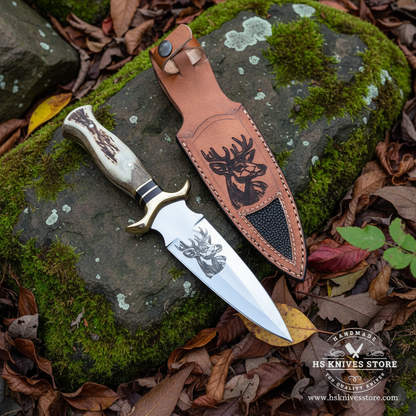 D2 steel dagger knife with stag antler handle and deer tooled leather sheath resting on mossy rocks in outdoor forest setting