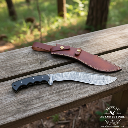 A fixed-blade knife with a patterned steel blade and black handle rests on a weathered wooden bench, with its brown leather sheath nearby. The background shows a blurred outdoor forest setting.